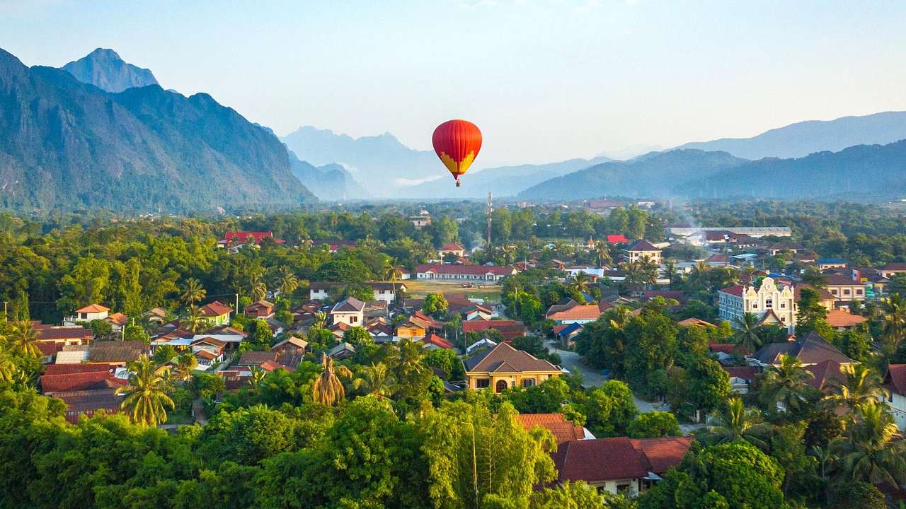 A hot air balloon over a town with lush greenery and houses, against hazy mountains