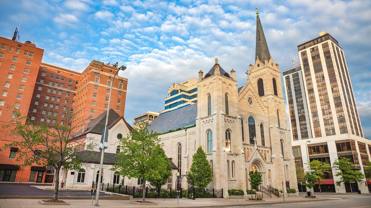 A church next to city buildings and trees under a blue sky with clouds