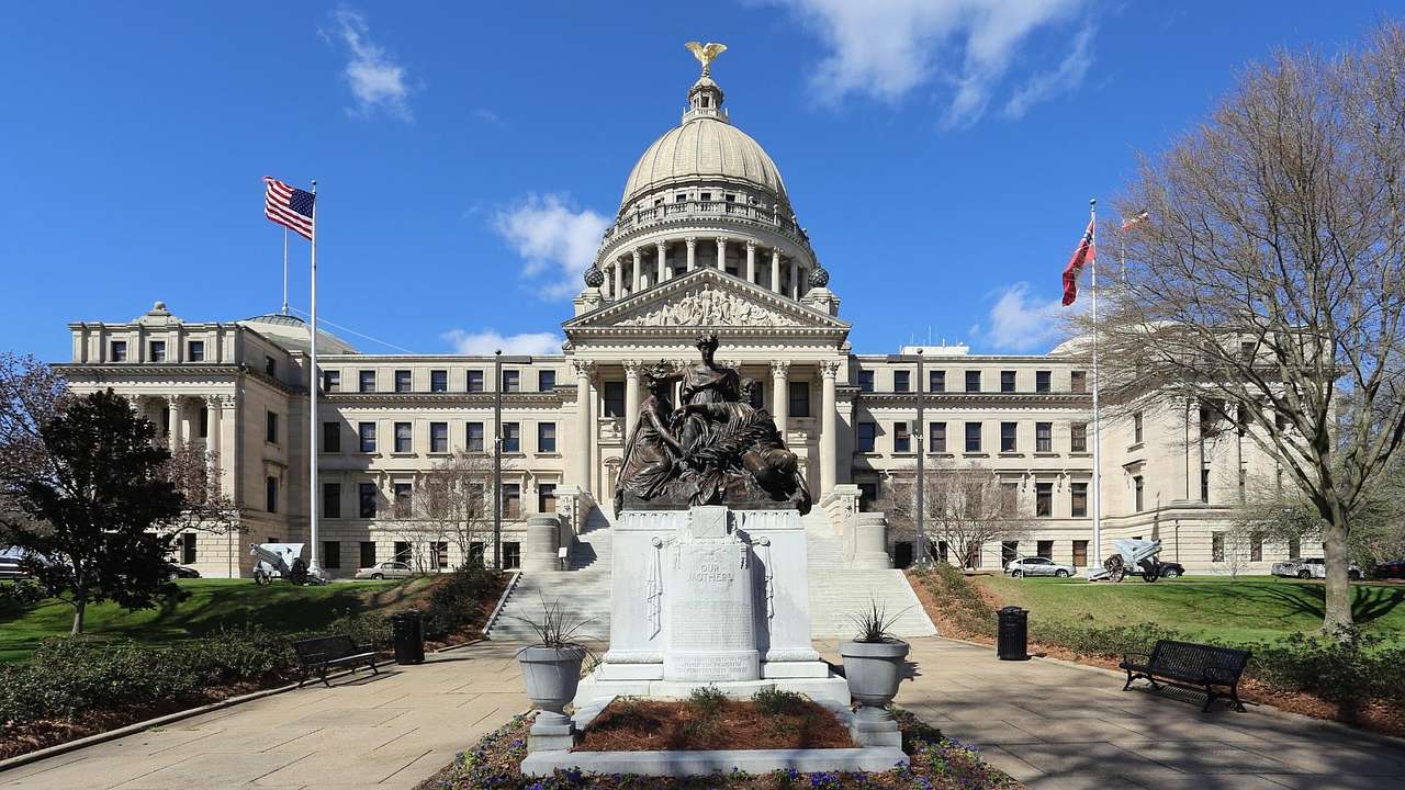 A state capitol building with a domed roof and a statue, flags, and trees in front