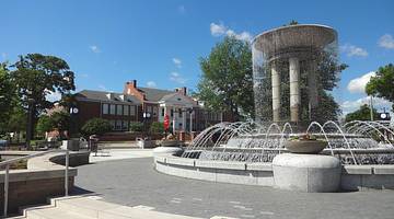 A large water fountain in a square with a red brick building and trees behind it