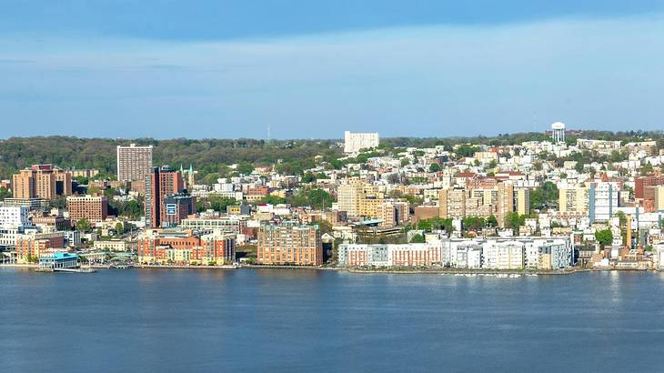 A city with many buildings and green trees behind it next to a lake under a blue sky