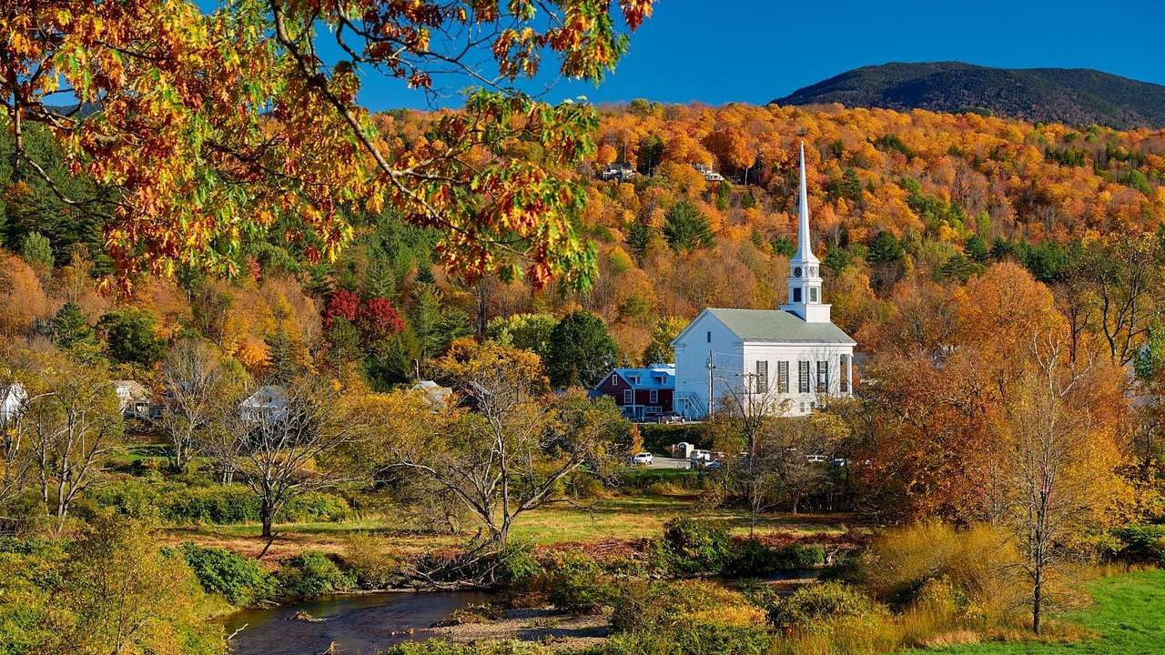 A white country church surrounded by fall foliage with a greenery-covered hill behind