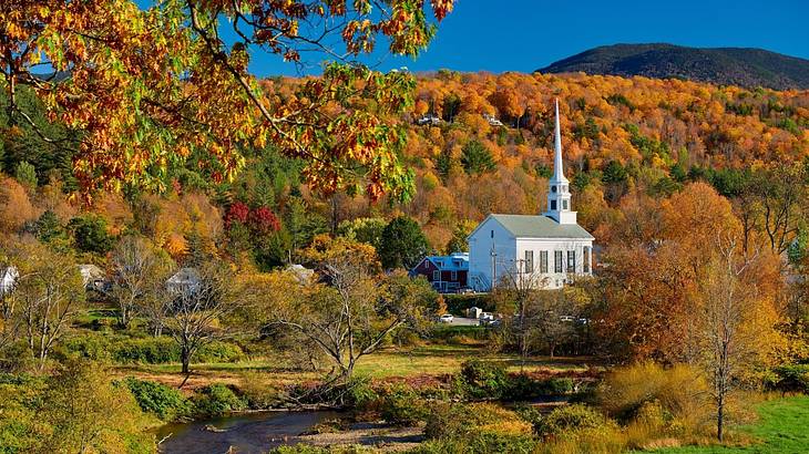 A white country church surrounded by fall foliage with a greenery-covered hill behind