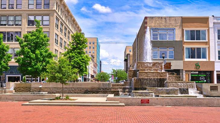 A red brick street with trees, buildings, and a water fountain behind it