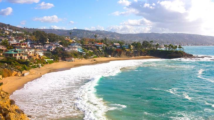 A beach with sandy shores and blue ocean, with a hillside town and greenery behind it
