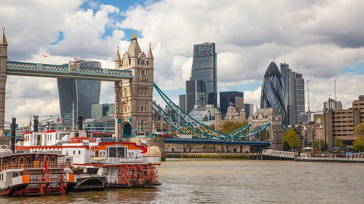 A paddle steamer on a river against a bridge & a cityscape under a mostly cloudy sky