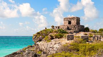 Ancient Mayan ruins on a rocky hill with green shrubs and the blue ocean behind