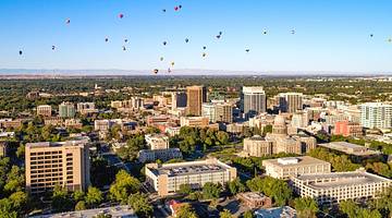 A cityscape with buildings and trees and colorful hot air balloons floating above it