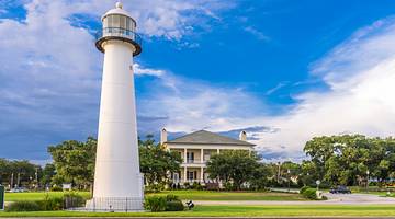A white lighthouse in front of a yellow and gray building surrounded by greenery