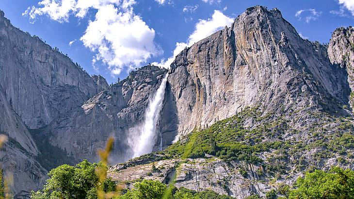 A tall rock formation with a waterfall with some greenery at the base on a sunny day