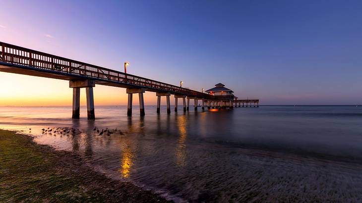 A boardwalk with yellow lights over water leading to a hut, under a clear evening sky