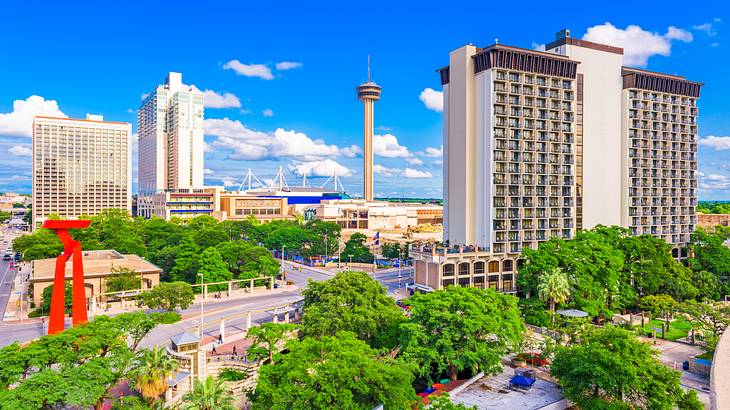 A city skyline with green trees in front of it under a blue sky with clouds