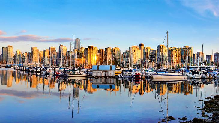 A panoramic skyline of tall buildings and boats reflected in still water at sunset