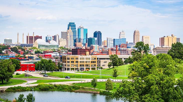 A park with green grass and trees and a pond and a city skyline behind it