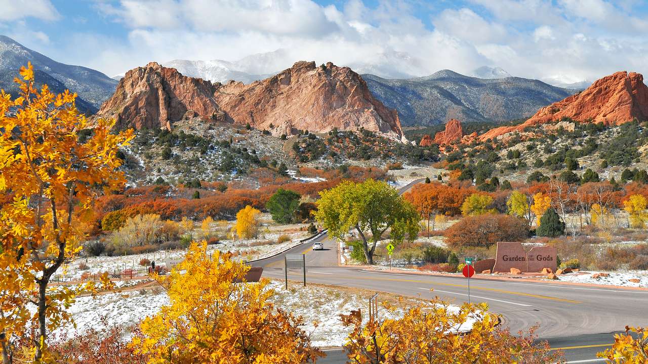A road with autumn-colored trees and light snowfall, with mountains in the background
