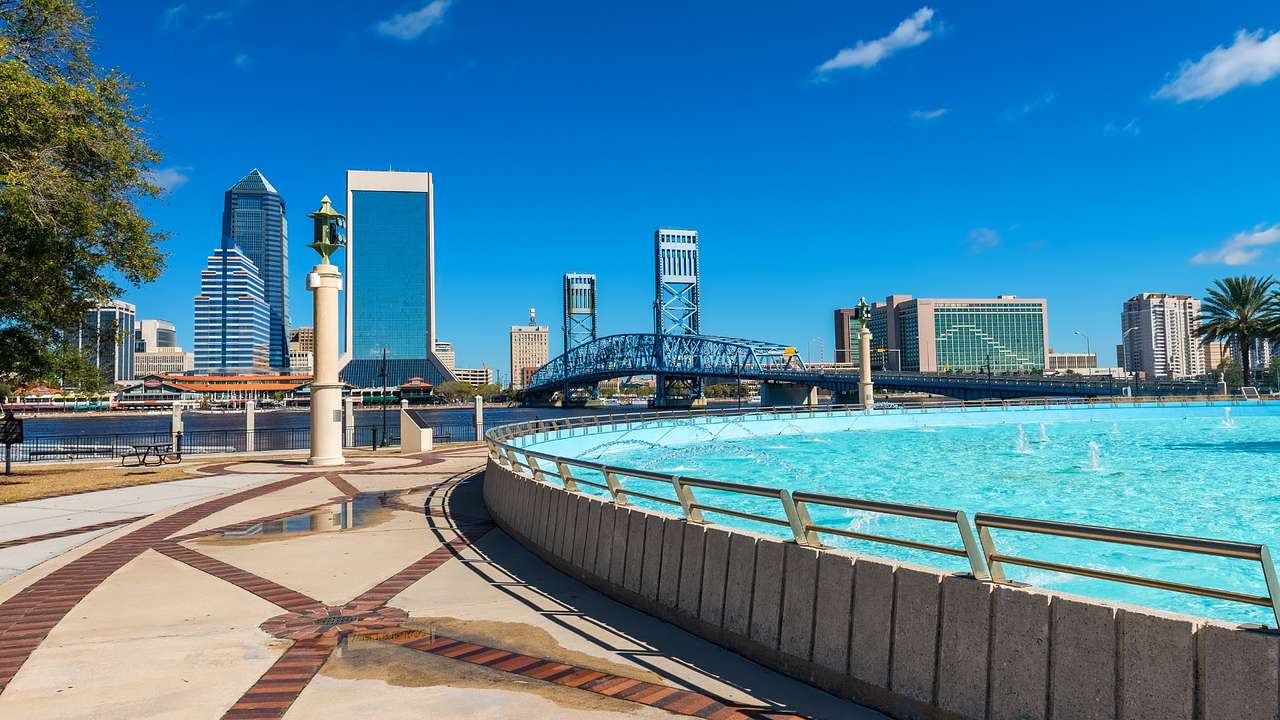 A path next to bright blue water with city buildings and a bridge at the back