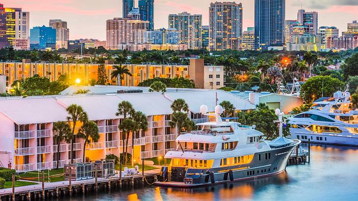 A view of a waterfront at sunset with boats, illuminated buildings, and trees