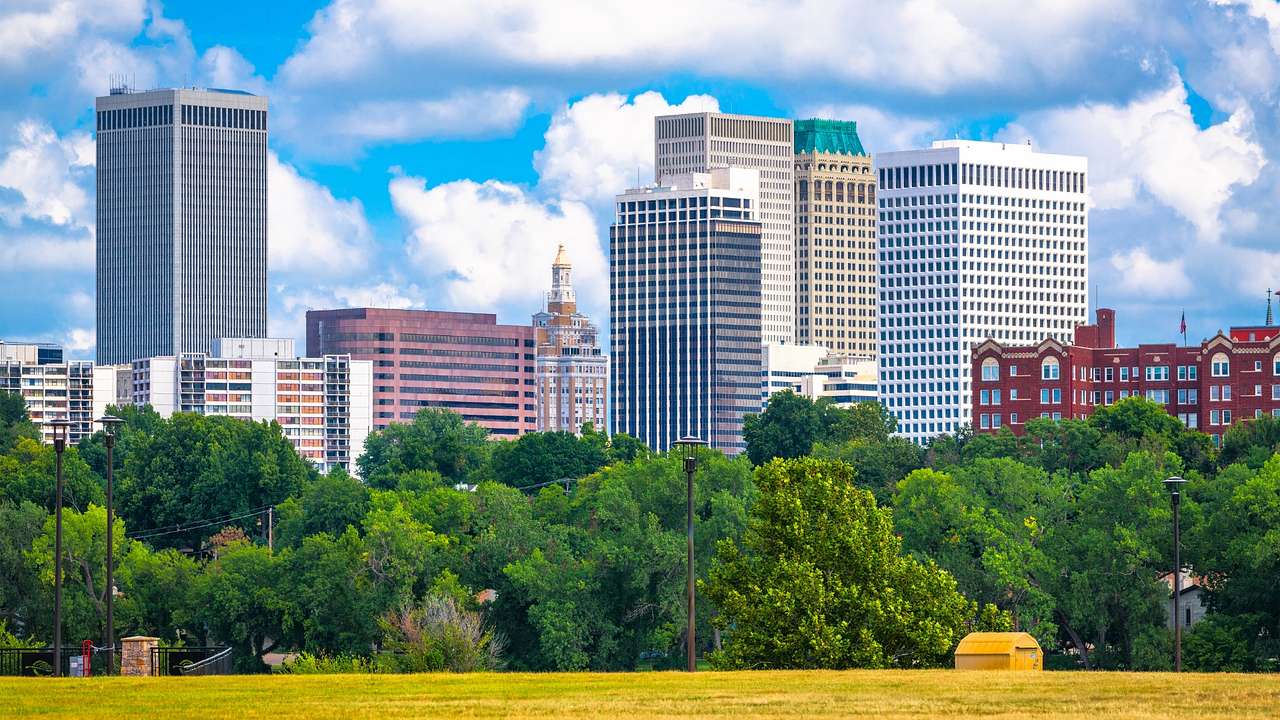 A city skyline next to green trees and grass under a blue sky with clouds