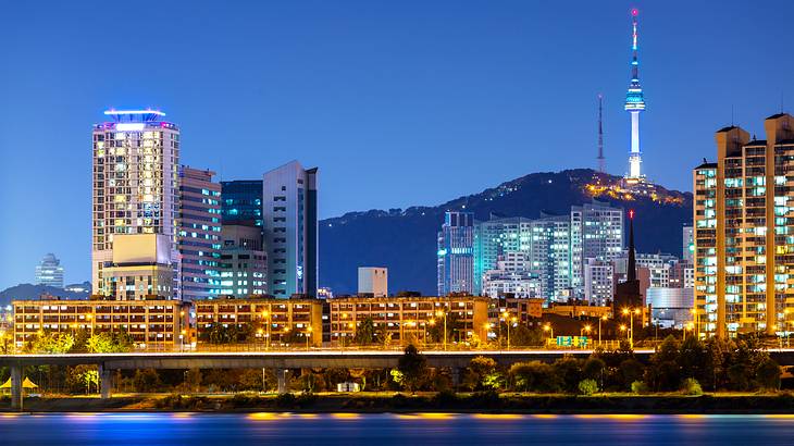 A city skyline at night with tall buildings lit up against a mountain in the distance