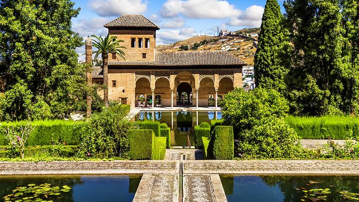 A yellow stone mansion surrounded by tall green trees with a water feature in front