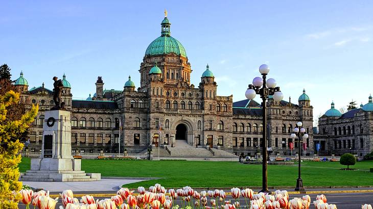 A grand historic building with green domes on top and greenery and flowers around it