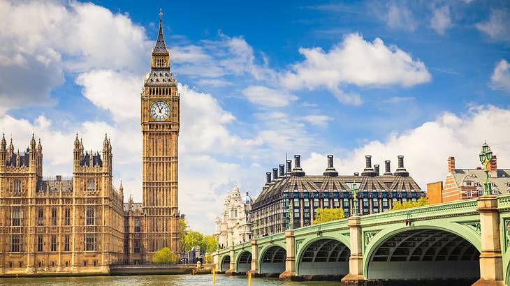 A clock tower next to buildings and a bridge over a river on a bright day