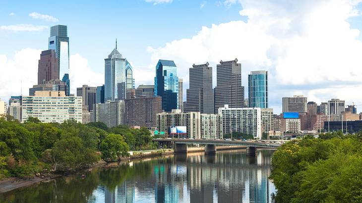 A city skyline with trees and a lake in front of it under a blue sky with some clouds