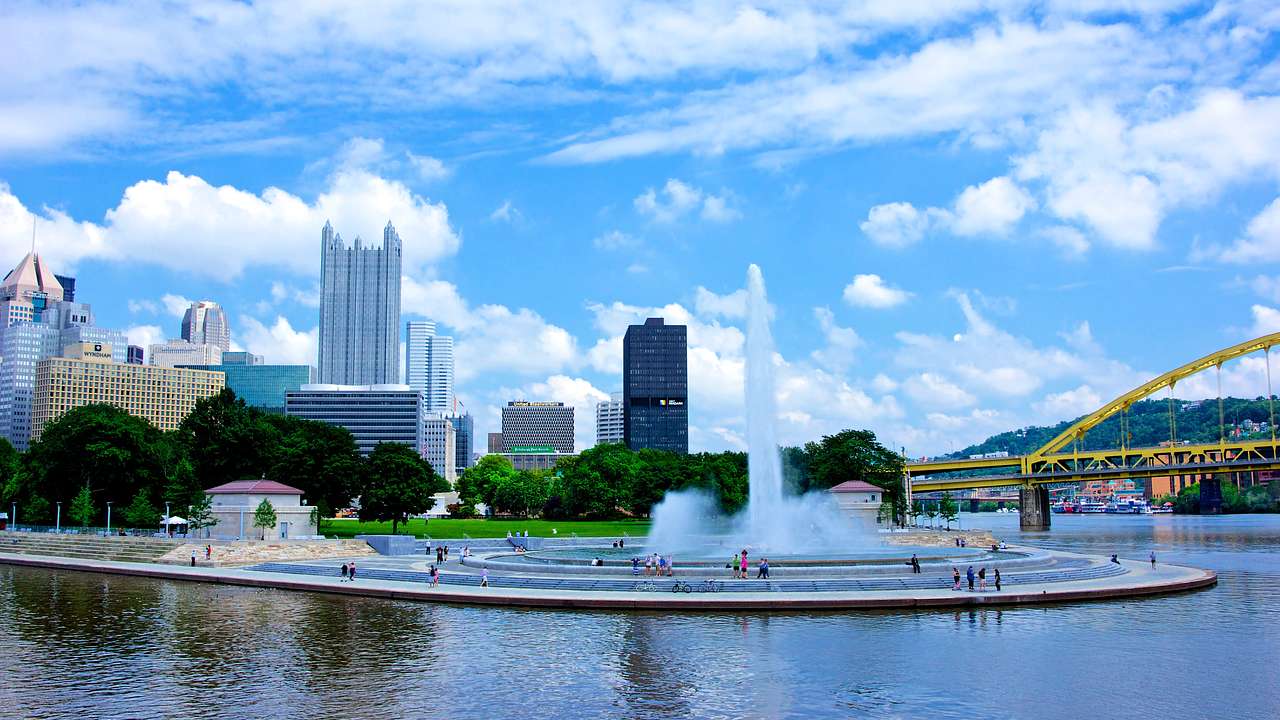 A fountain near a bridge and a body of water with the city skyline in the background