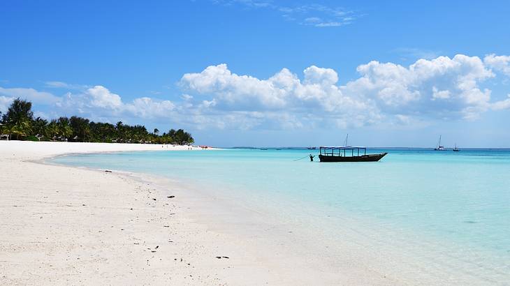A white sand beach next to trees and the blue ocean with a boat on it