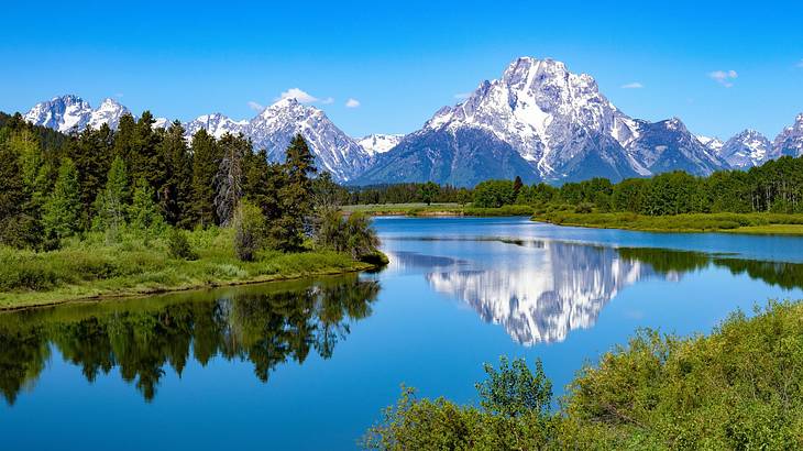 Snow-capped mountains next to a river and green trees under a blue sky