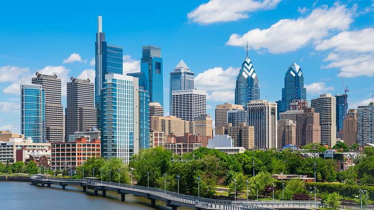 A city skyline next to the water and trees under a bright blue sky with clouds