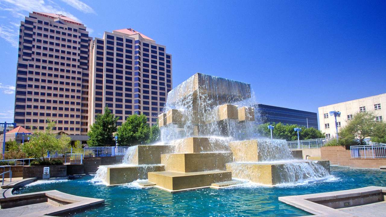 A water fountain near buildings and trees under a blue sky