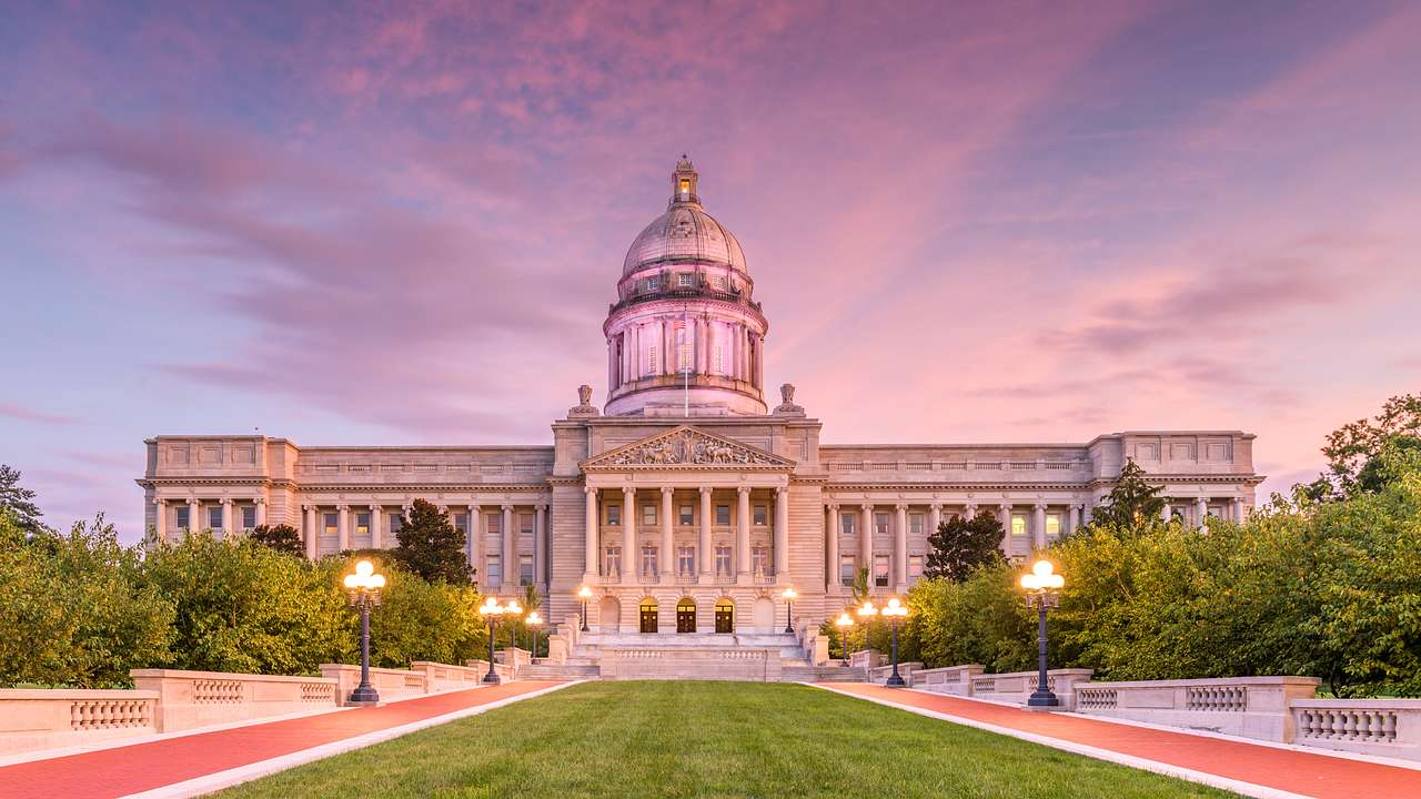 A state capitol building with a dome roof and a green lawn in front, at sunset