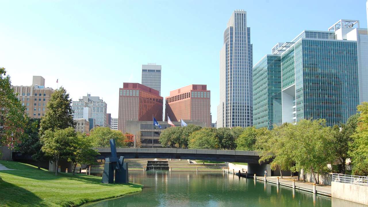 A body of water surrounded by grass and trees with a bridge and buildings in the back