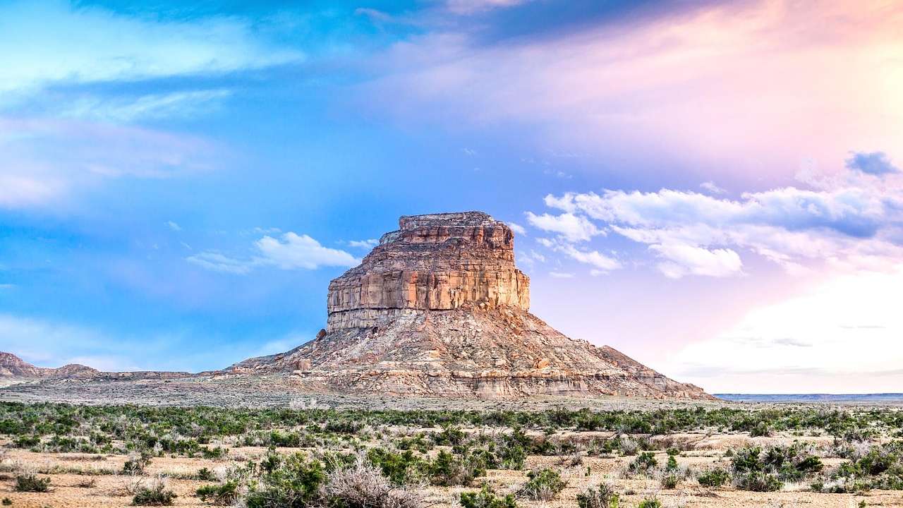 A butte on an empty field with small bushes, under a partly cloudy purple sky