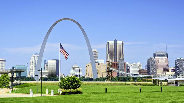 A city skyline with skyscrapers next to a tall arch, grass, and an American flag