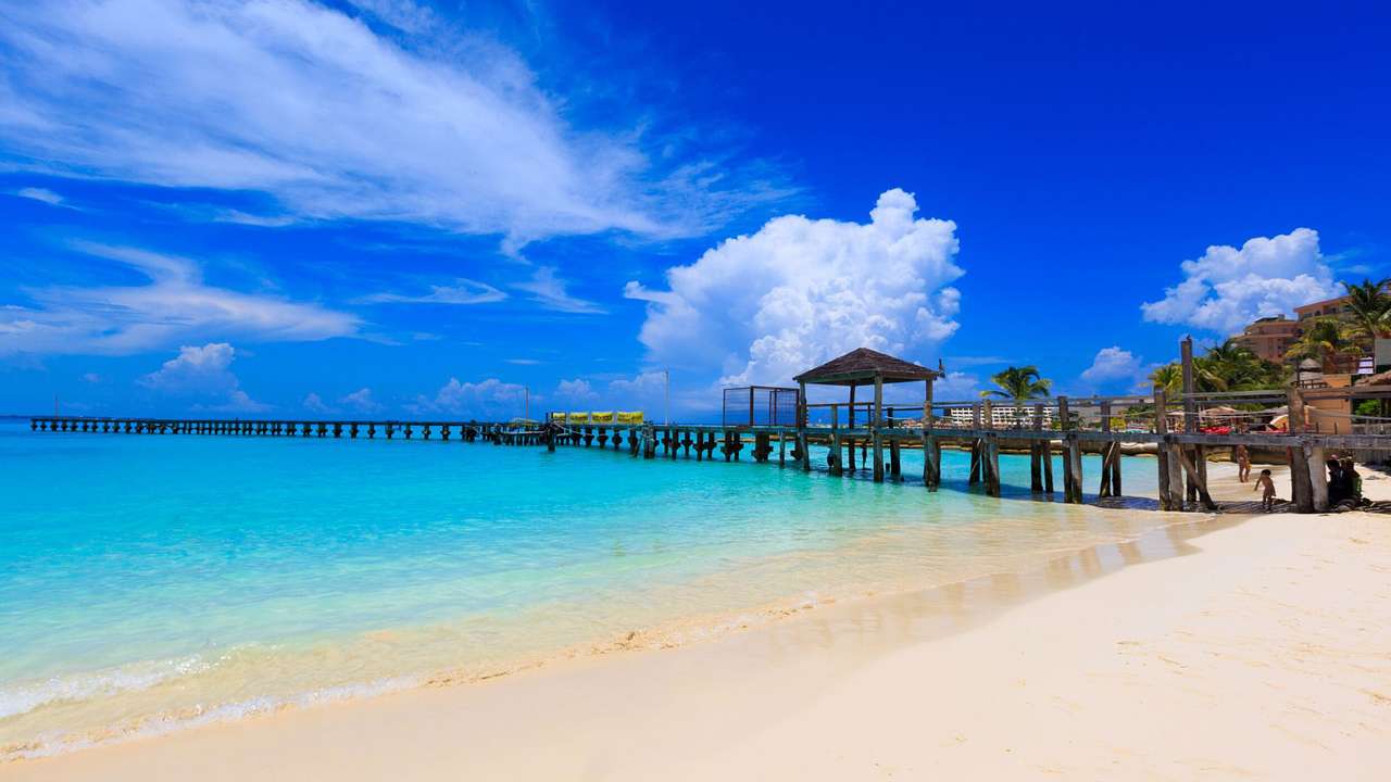A white sand beach next to blue water and a small pier under a blue sky