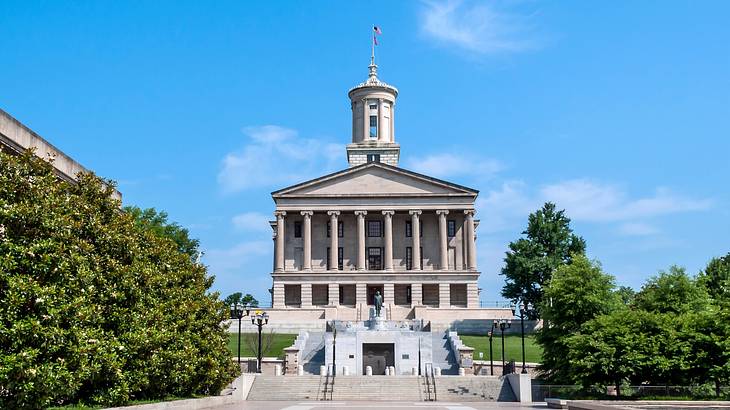 A large stone building with columns next to stairs, green trees, and a blue sky