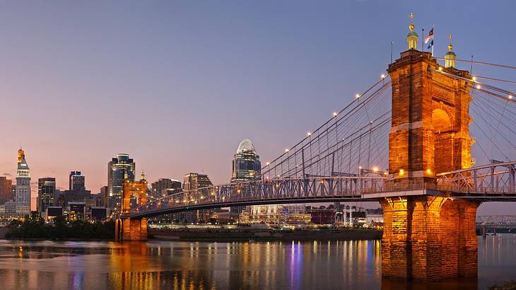 A bridge illuminated at night with water under it and a city skyline on the shore