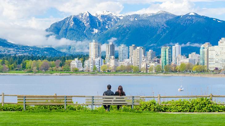 A couple sitting on a bench on the grass next to water, a city skyline, and mountains