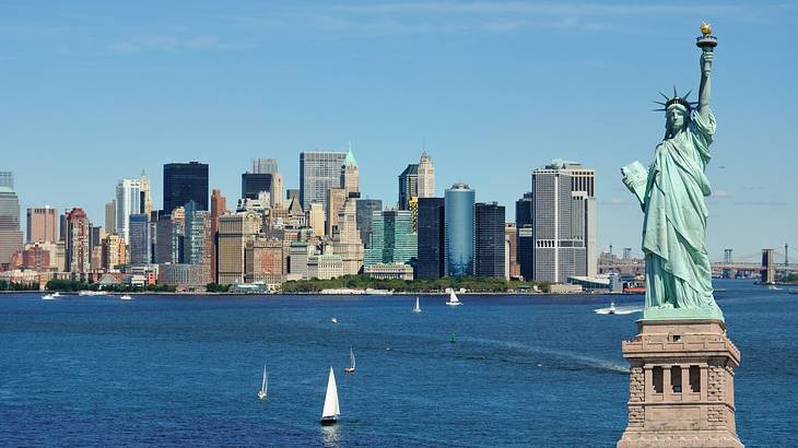 The blue Statue of Liberty and tall buildings and water in the background