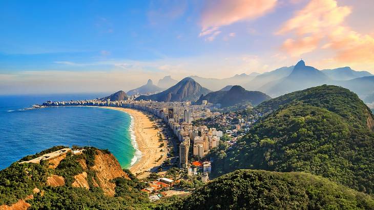 A sandy beach with blue water and city buildings surrounded by lush mountains