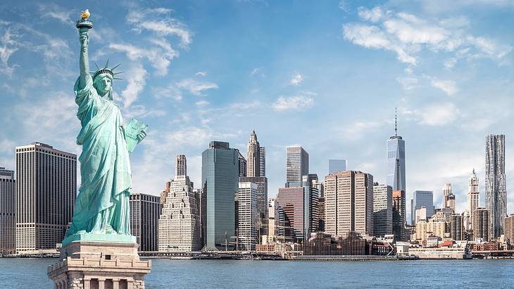 The blue Statue of Liberty and tall buildings and water in the background