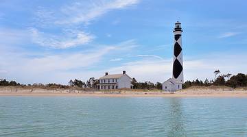 A beautiful sandy beach with blue water and a white house and lighthouse on the shore