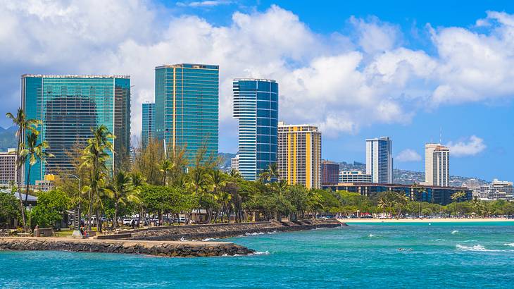 Mirrored skyscrapers with palm trees and the ocean in front of them under a blue sky