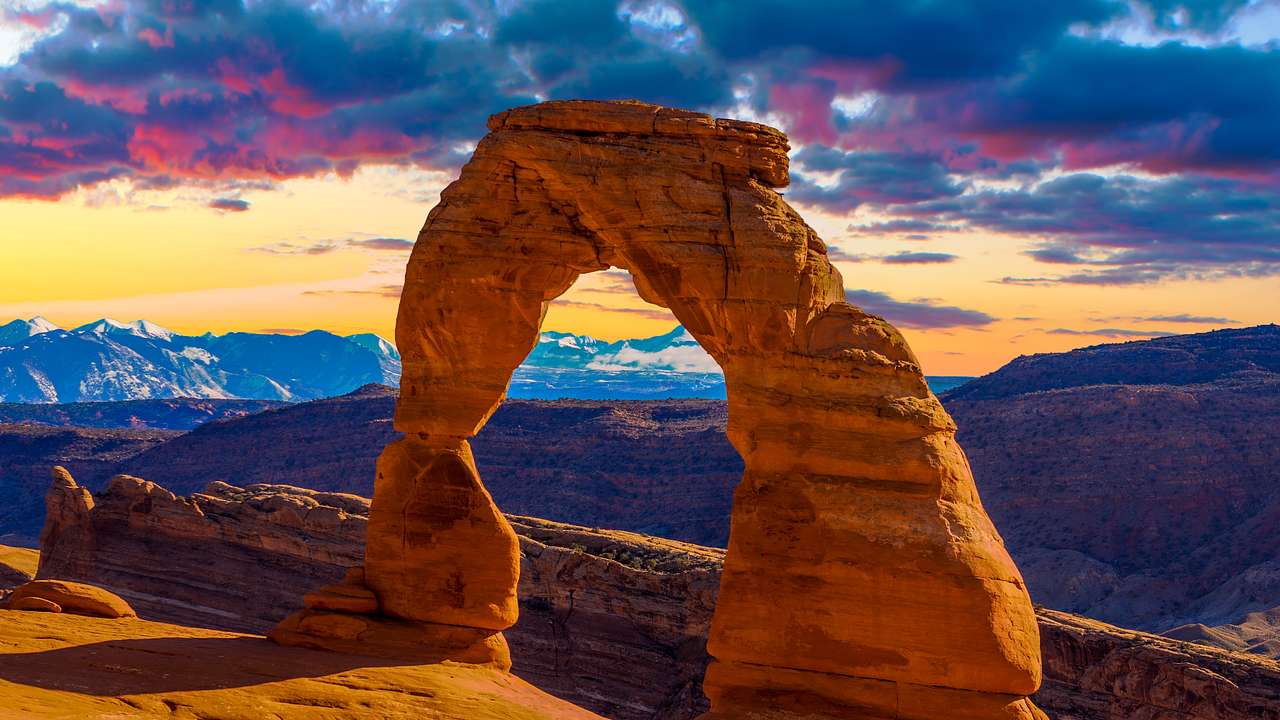A stunning red-hued sandstone arch atop a cliff with mountains behind
