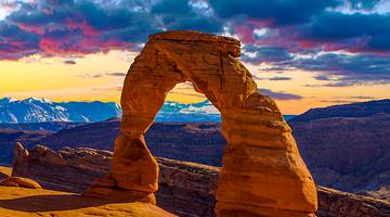 A stunning red-hued sandstone arch atop a cliff with mountains behind