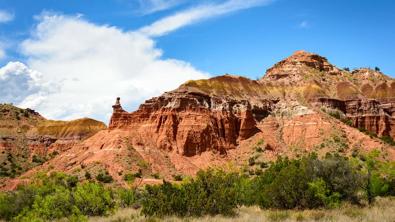 A large red barren rock face from below, with dry green shrubbery in front