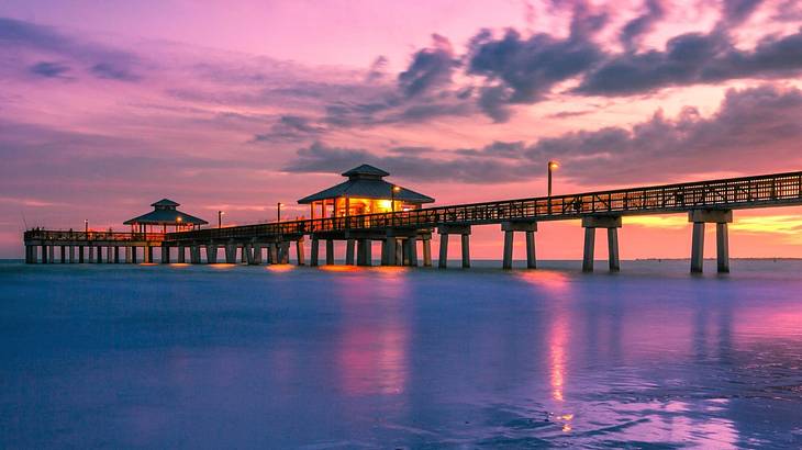 A boardwalk with 2 huts over water against a sunset with a partly cloudy purple sky