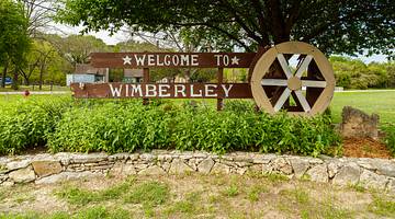 A "welcome to Wimberley" sign with a wheel on the right, surrounded by greenery